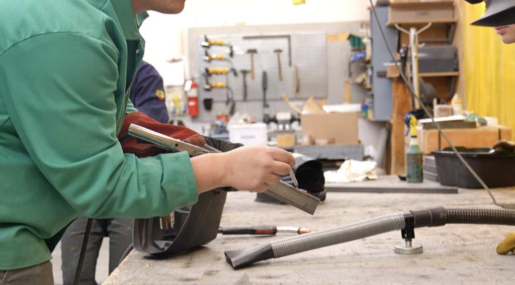 Connor Thompson points at a steel tube showing students what makes a good weld